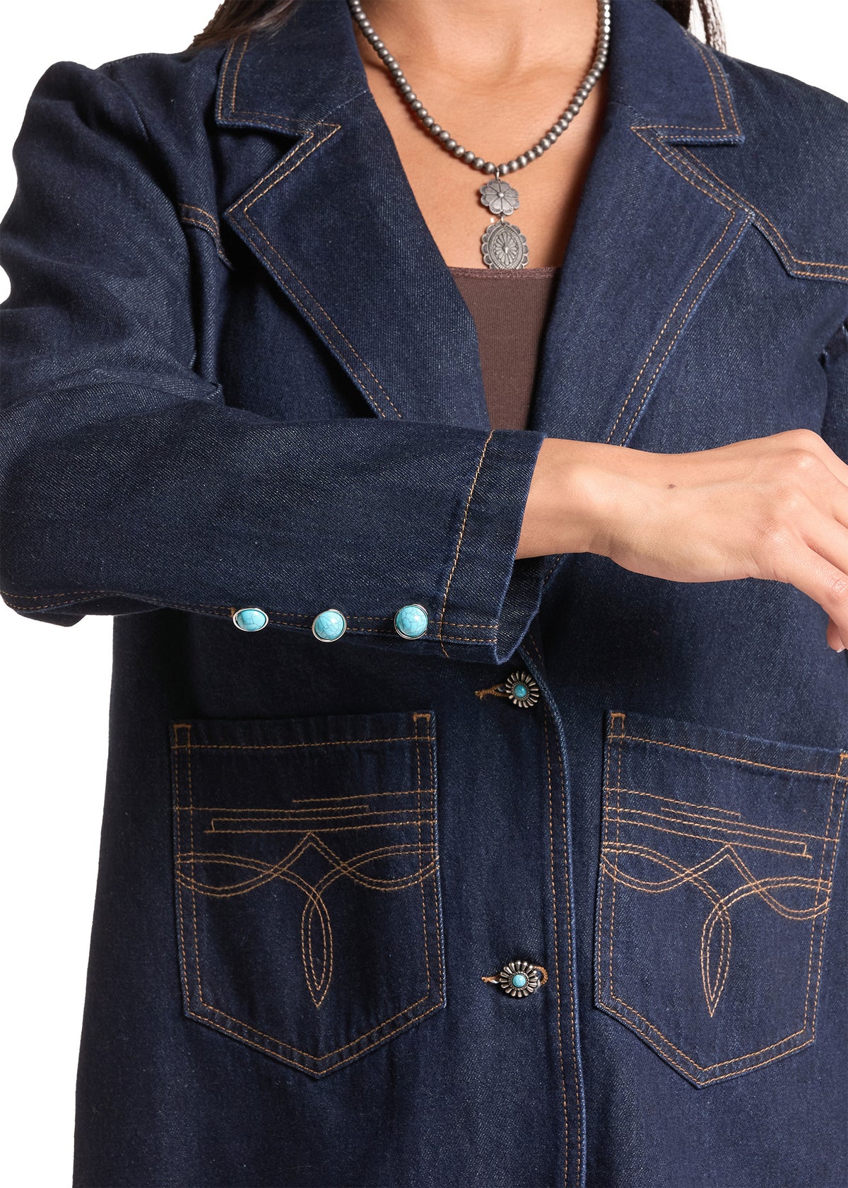 A close-up of a dark blue denim jacket with decorative gold contrast stitching, turquoise-colored buttons on the cuff, and matching turquoise accents on the front pockets and buttons. The jacket is shown on a model wearing a brown top and a silver pendant necklace.