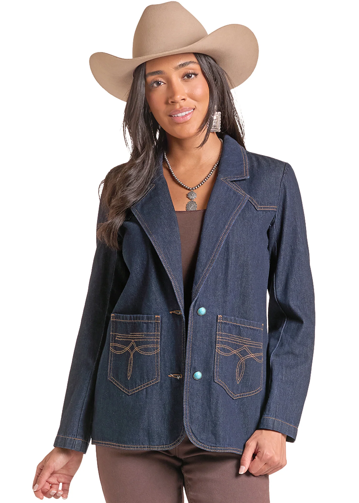 A woman modeling a dark blue denim western-style blazer with contrast tan stitching, decorative front pockets, and turquoise buttons, paired with a tan cowboy hat, brown top, and brown pants against a white background.