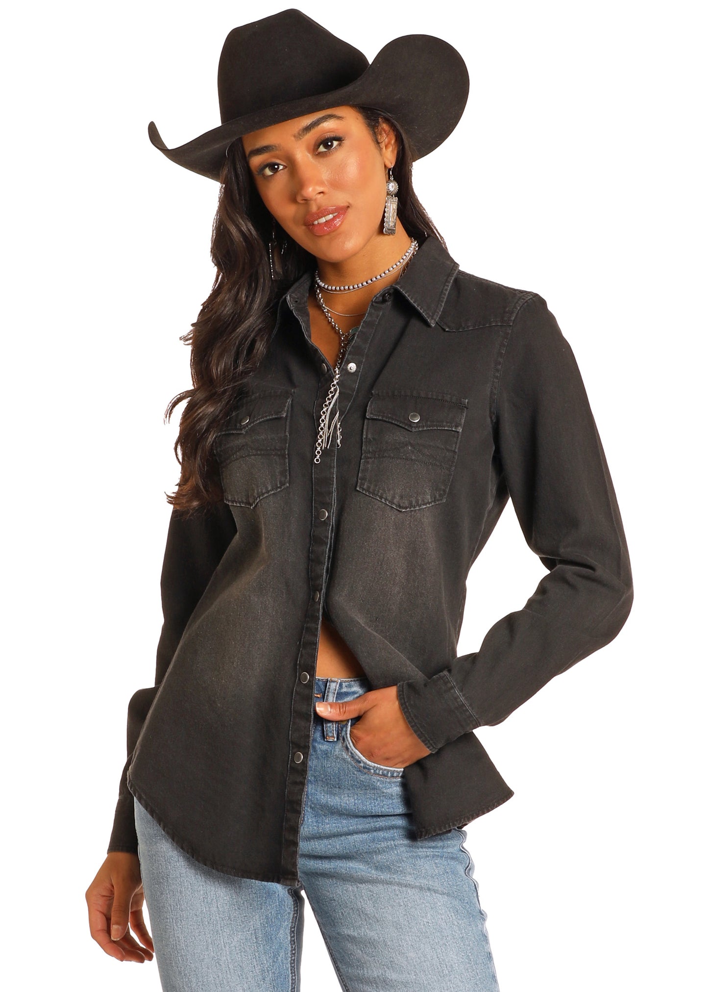 A cowgirl poses against a white background wearing the Rock & Roll Denim Black Denim Long Sleeve Shirt, blue jeans, a black cowboy hat, and silver jewelry, with one hand in her pocket.