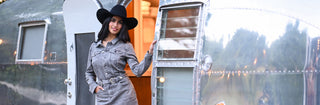 A woman wearing a gray denim dress and a black wide-brim hat stands in the doorway of a silver Airstream trailer.