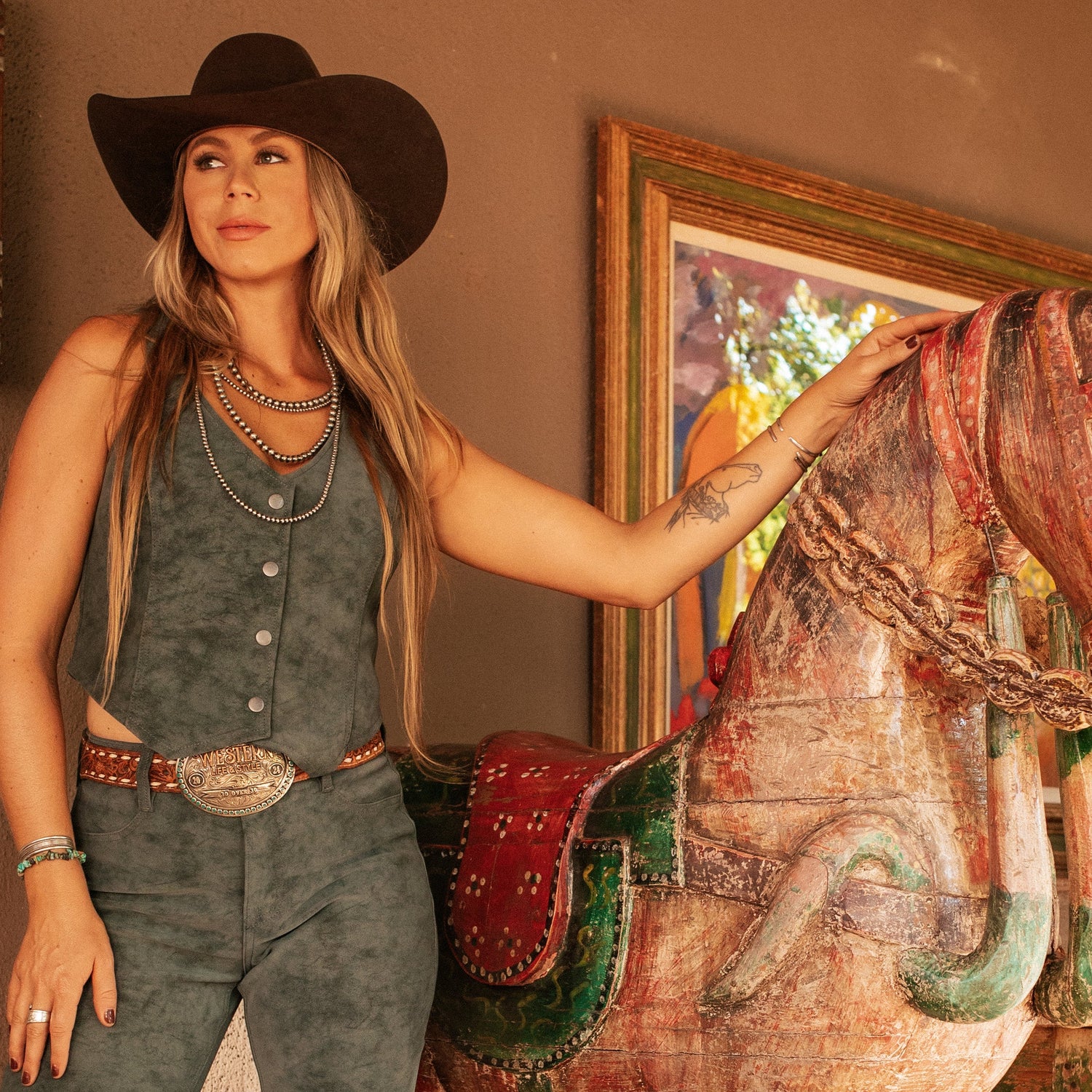 Woman in cowboy hat and outfit standing next to a decorative horse figure indoors.