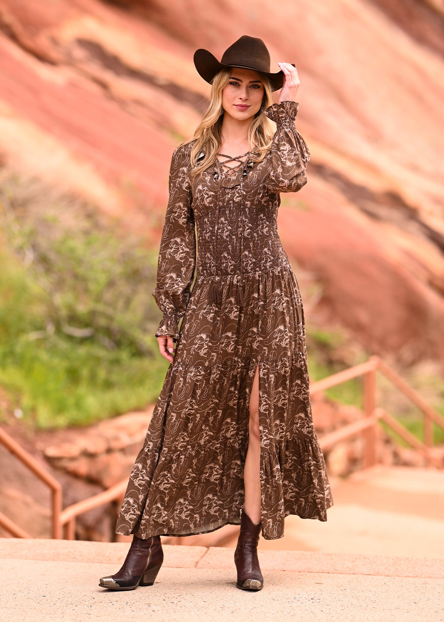 A woman in a brown patterned Rock & Roll Denim Challis Peasant Smocked Maxi Dress with ruffle details, a black cowboy hat, and ankle boots stands on a path by red rock formations, gently holding her hat and looking at the camera.