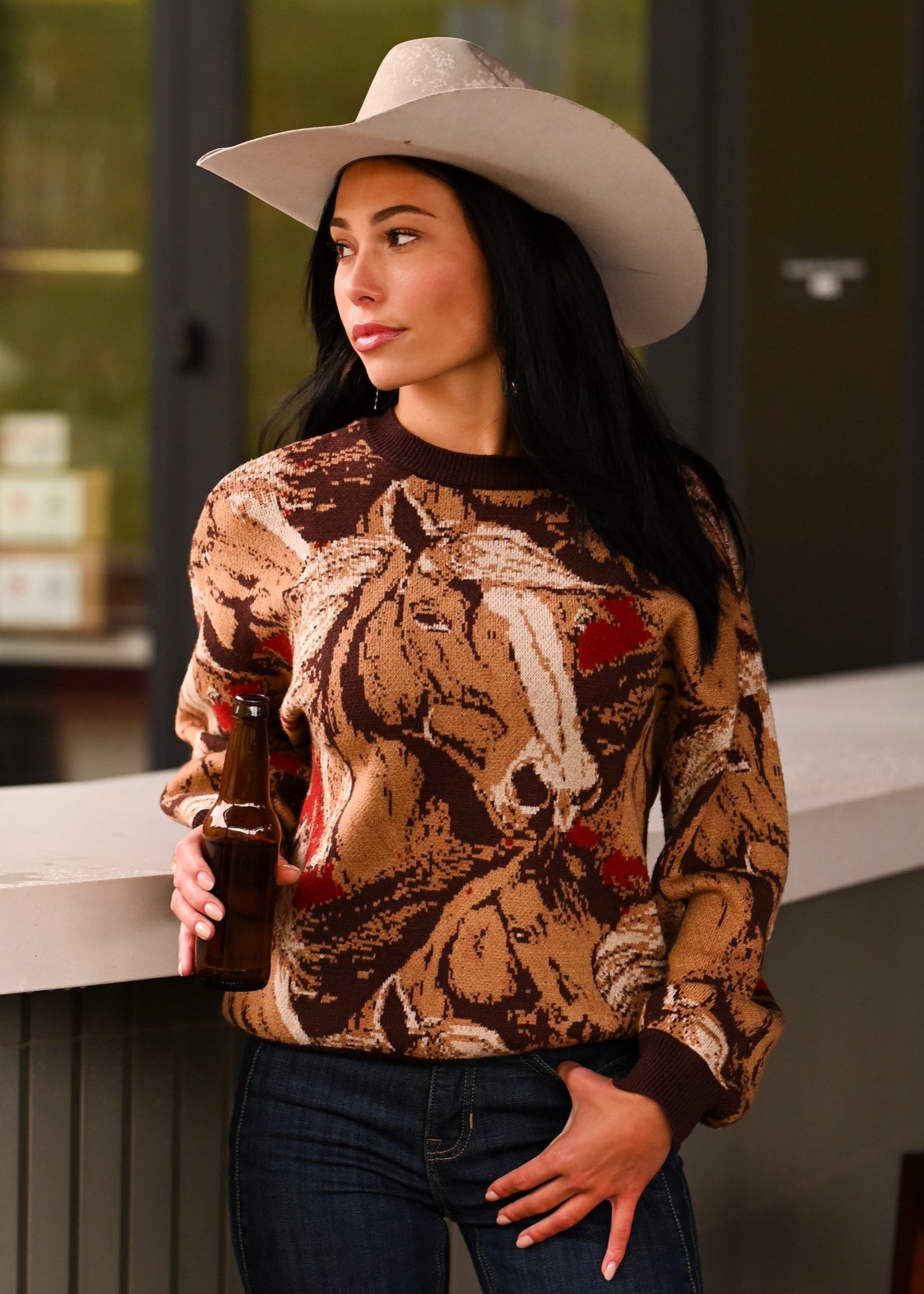 A woman in a Rock & Roll Denim Oversized Horse Sweater, cowboy hat, and jeans stands by a counter holding a brown bottle, gazing to the side.