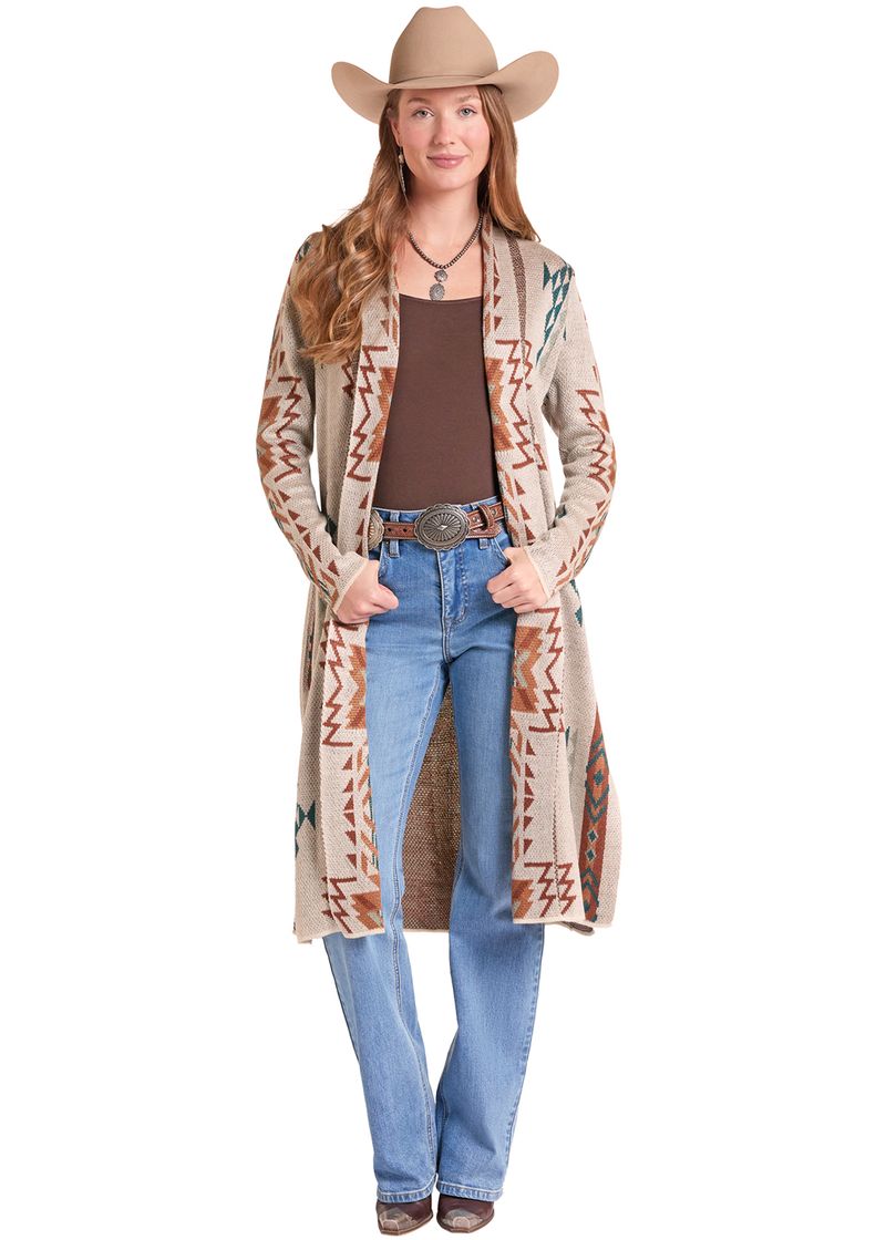 A woman modeling a long beige cardigan with brown, orange, and blue geometric patterns, styled with a brown tank top, blue jeans, a western belt, and a tan cowboy hat against a white background.
