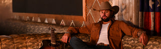 Koe Wetzel sitting on a couch wearing a cowboy hat, an unbuttoned brown shirt and denim. 