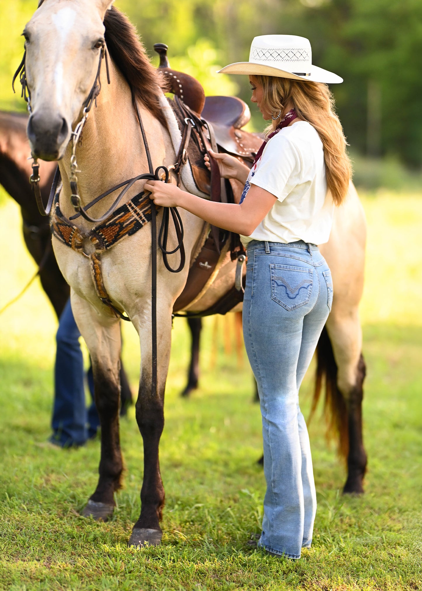 A woman in a white cowboy hat, white t-shirt, and light blue jeans stands on a grassy field next to a light brown horse wearing a leather saddle and bridle.