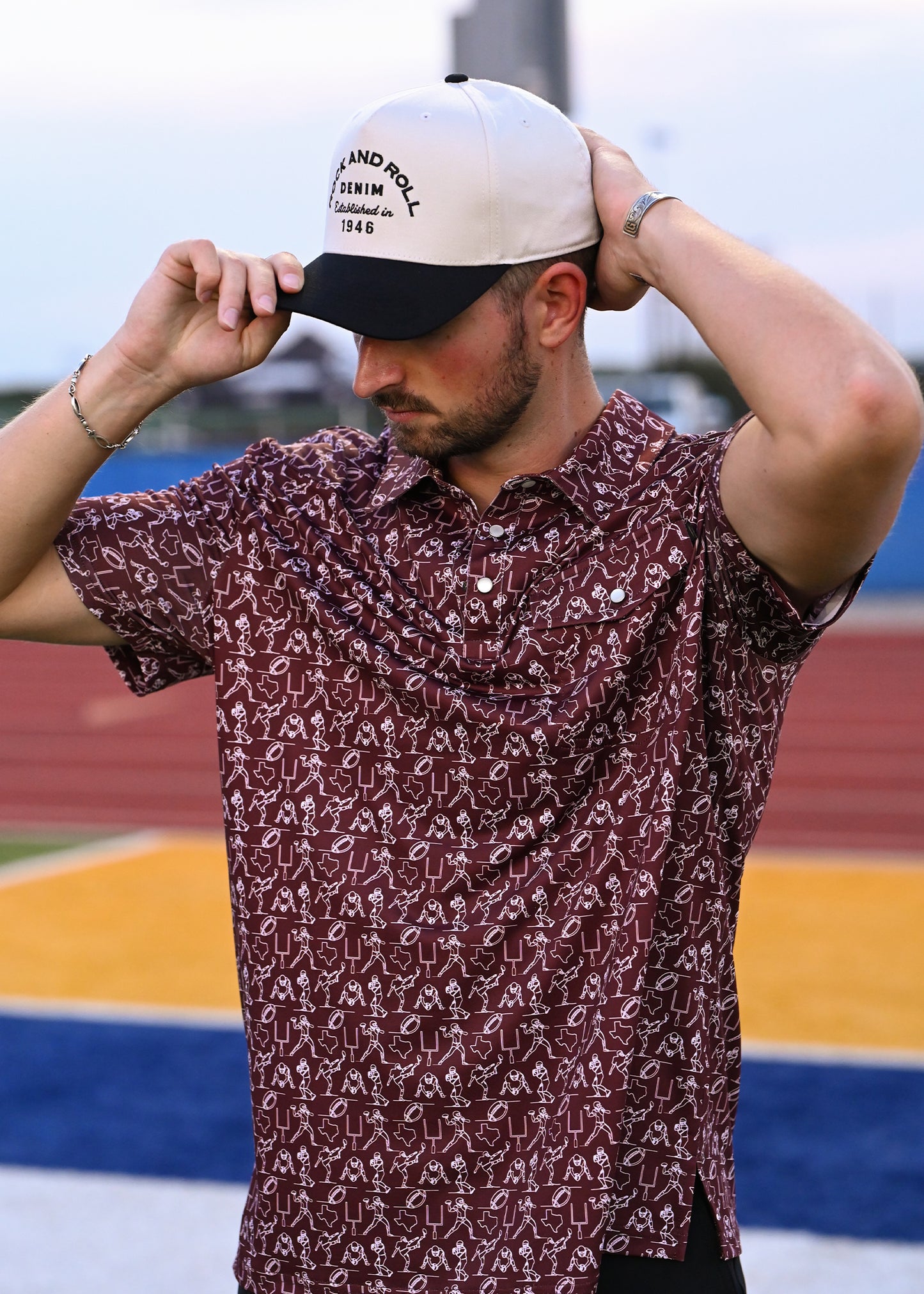 Man wearing adjusting his hat while wearing a maroon and white polo on a football field. 