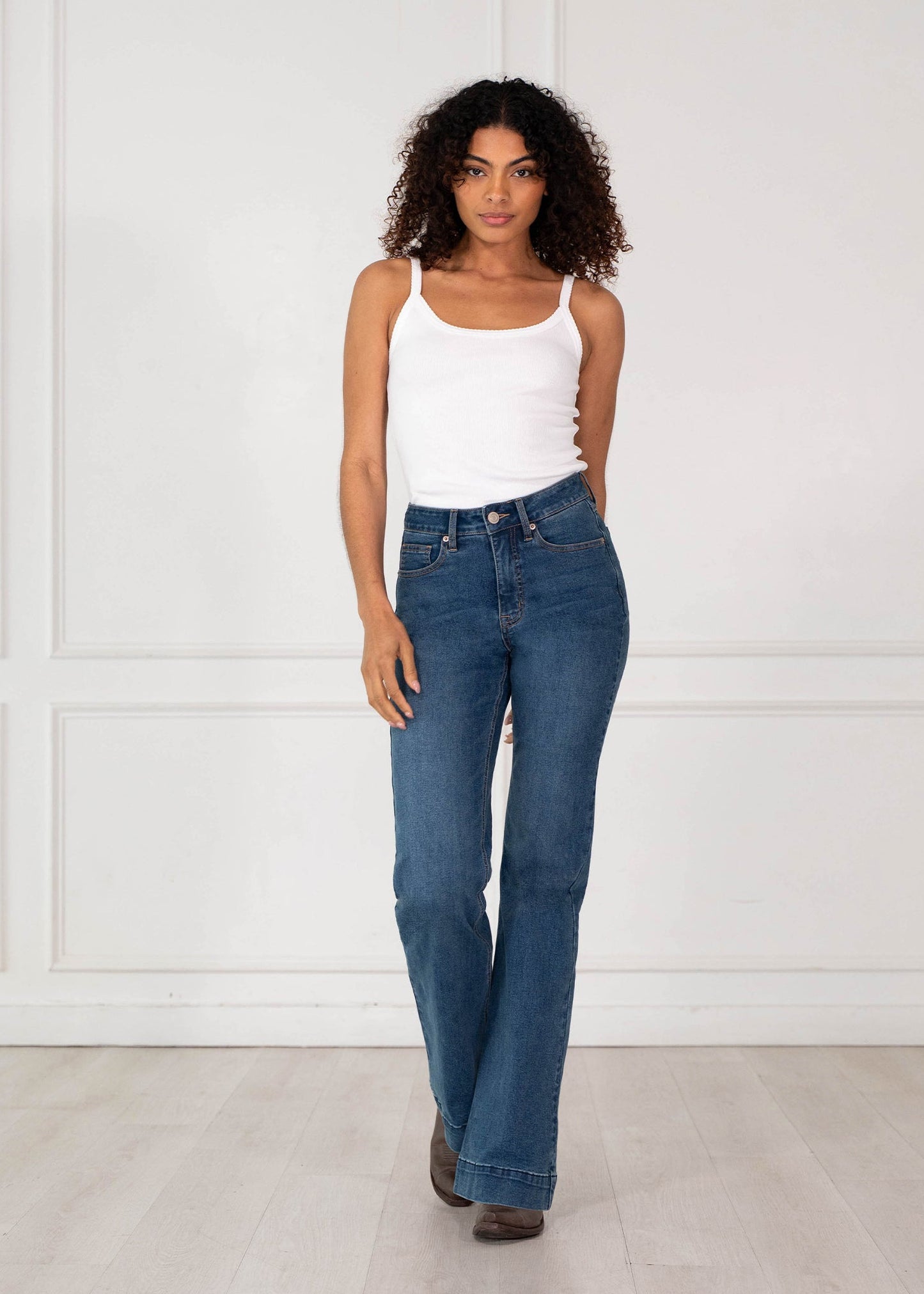 A woman with curly dark hair stands in a bright room wearing blue denim jeans, a white tank top, and brown shoes.
