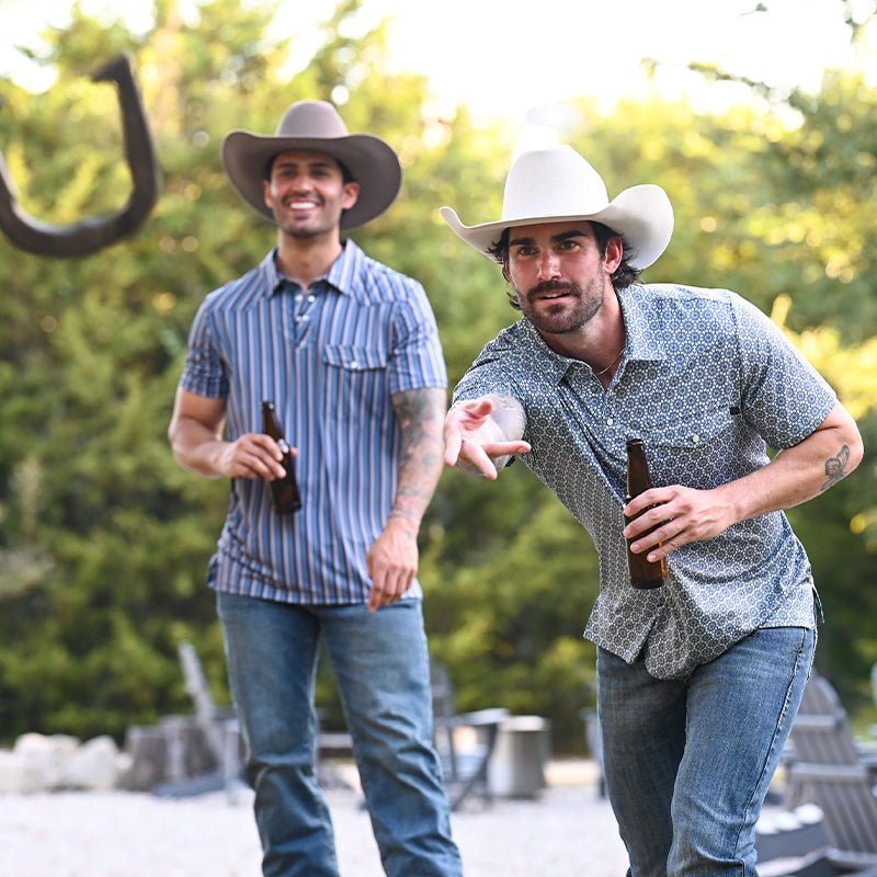 Two men playing horseshoes outside, drinking beer and wearing cowboy hats, polos and jeans. 