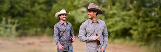 Two men wearing western-style button-down shirts and cowboy hats walking outdoors in a wooded setting.