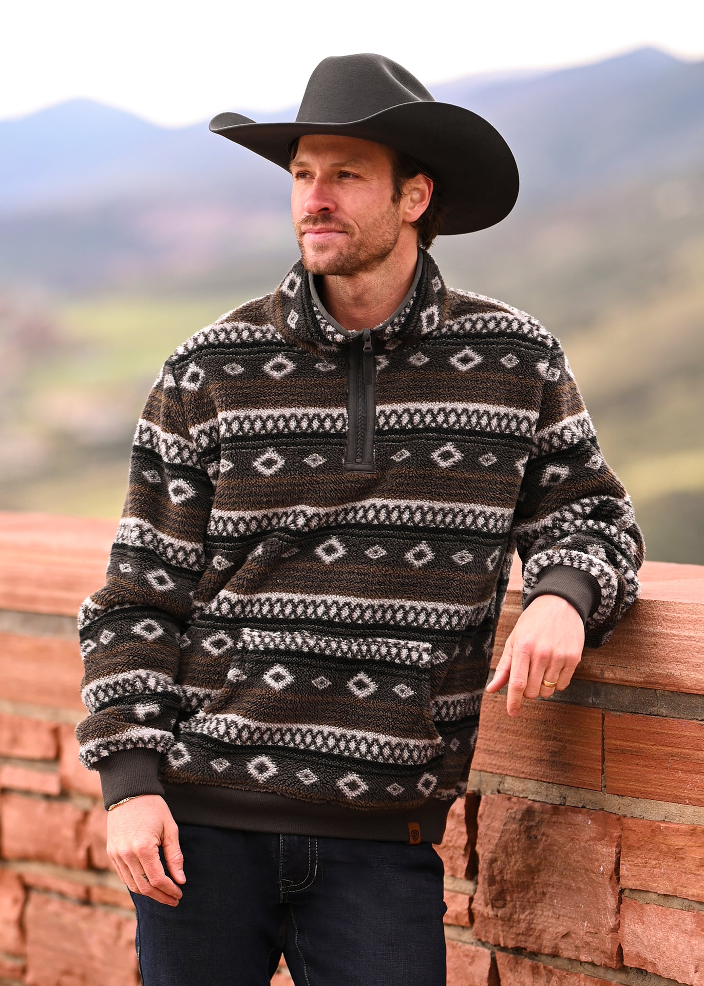 A man in a black cowboy hat and Rock & Roll Denim's Chocolate Aztec Print Fleece Pullover leans against a brick wall outdoors, with blurred mountains and greenery in the background.