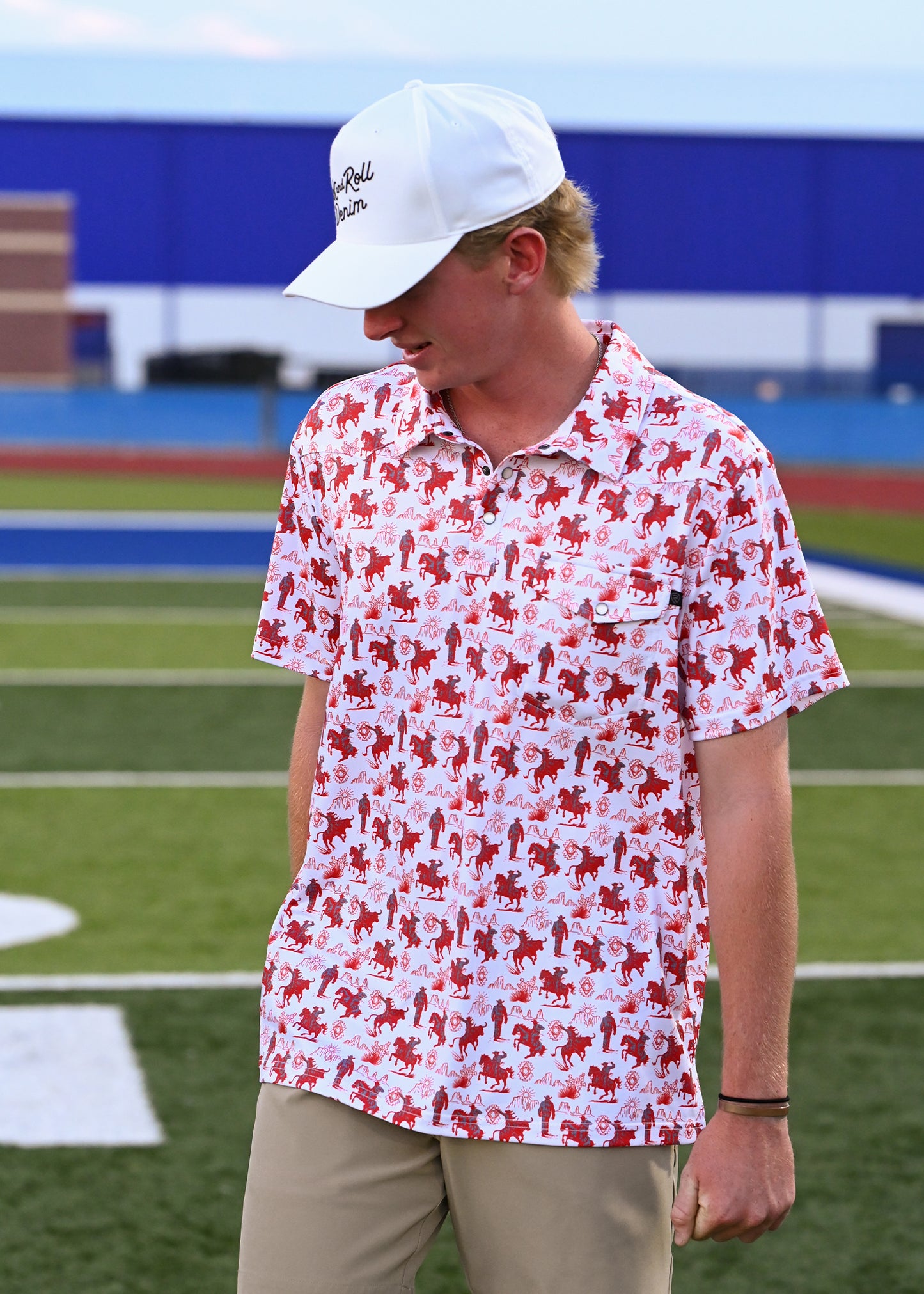 A man wearing a white baseball cap and a white short-sleeve polo with a red and white dog print, standing on a green athletic field.