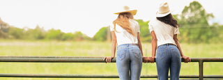Two women standing side by side from behind, wearing cowboy hats, white t-shirts, and blue jeans, leaning on a metal fence in an outdoor field.
