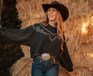 Woman in a black embroidered shirt and cowboy hat standing in front of hay bales.