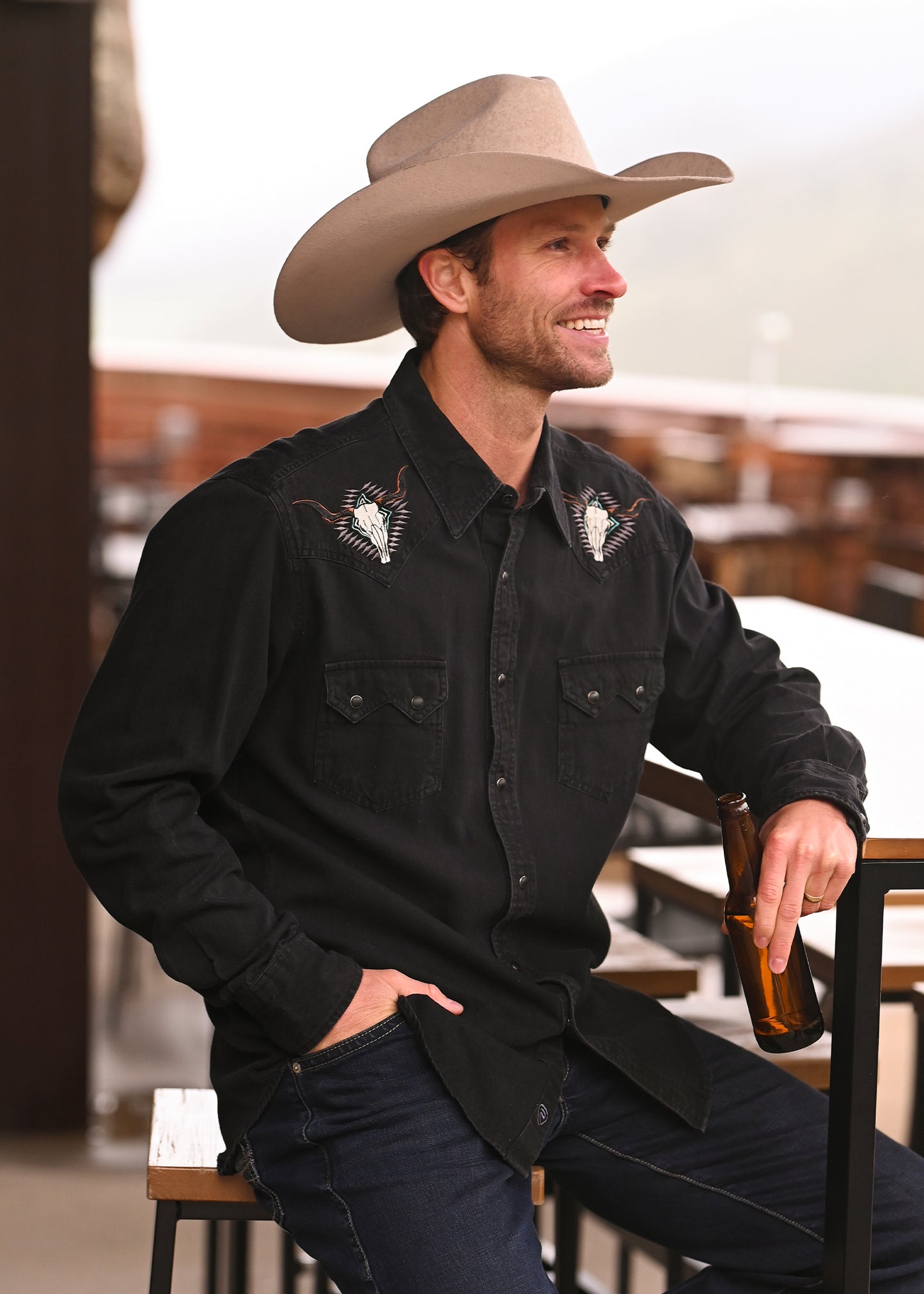 A man in a Rock & Roll Denim Slim Fit Embroidered Black Denim Snap Shirt and tan cowboy hat sits on a stool outdoors, smiling and holding a brown glass bottle.