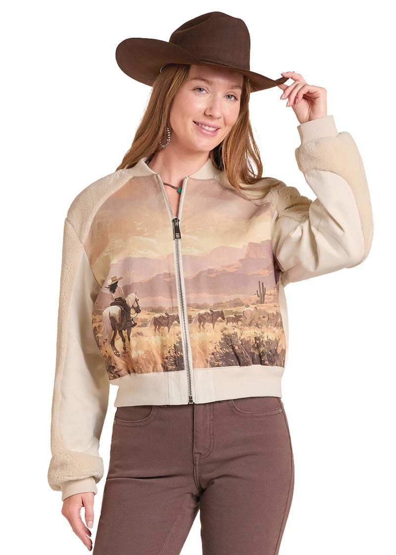 A woman wearing a cropped zip-up bomber jacket with a western desert landscape print on the front and beige sherpa sleeves, paired with brown pants and a brown cowboy hat against a white background.